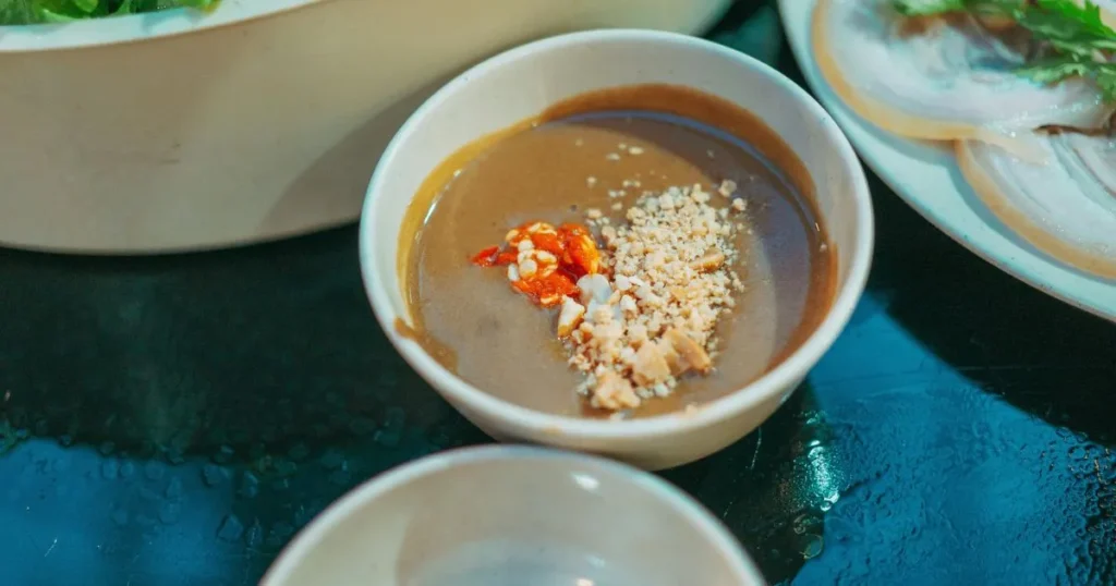 Homemade peanut sauce in a bowl placed on a kitchen counter, with a spoon and fresh ingredients nearby.