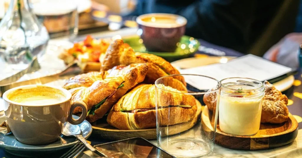 A cappuccino beside buttery pastries topped with powdered sugar and strawberries in a cozy kitchen setting.